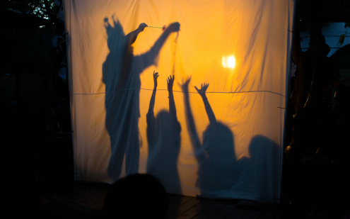 The silhouette of three children is shown on an orange backlit sheet. The children have their hands in the air and are reaching up towards the light.