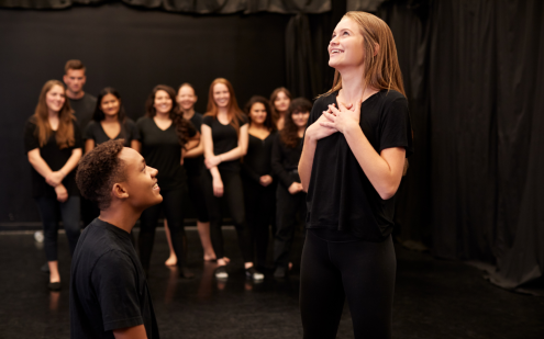 A group of teenagers are stood in a studio space, all wearing all black. A couple are in the forefront acting out a scene, whilst a group behind observe.